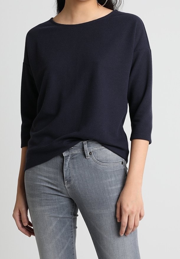 Mörkblå, texturerad stickad tröja med vid halsringning och ¾-långa ärmar, kombinerad med ljusgrå jeans med klassisk passform och subtila detaljer.