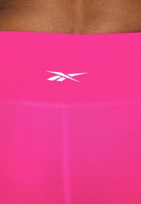 Růžové sportovní legíny s přiléhavým střihem a širokým pasem s bílým logem, vyrobené z hladkého, elastického materiálu s jemnými švy.
