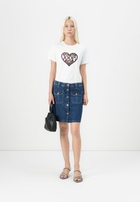 Tricou alb cu grafică în formă de inimă și text, asortat cu o fustă creion din denim albastru, cu nasturi în față și două buzunare aplicate. Geantă de umăr neagră și sandale cu curele.
