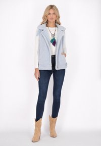 Himmelblauer, flauschiger Weste mit Kapuze, kombiniert mit einem weißen Langarmshirt und dunklen Jeans. Nussfarbene Wildleder-Stiefeletten runden den Look ab.