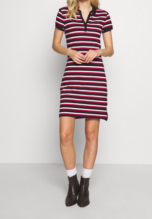 Kobieta ubrana w czarną, białą i czerwoną sukienkę z krótkim rękawem w poziome paski z kołnierzykiem, białe skarpetki i czarne botki za kostkę, stojąca nieruchomo.