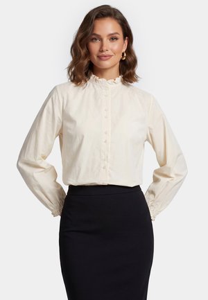 Vrouw met golvend bruin haar draagt gouden oorbellen in de vorm van ringen, een crèmekleurige blouse met ruches aan de kraag en een zwarte rok met hoge taille tegen een effen witte achtergrond.