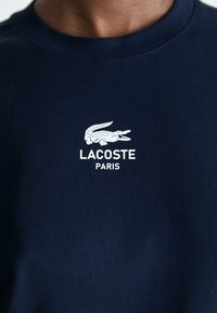 Navy blue T-shirt made of cotton, featuring a white embroidered crocodile logo and "LACOSTE PARIS" text below. Rounded neckline.