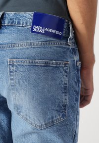 Šviesiai mėlynos džinsinės kelnės su nublukusia tekstūra ir kontrastingais siūlais. Ant juosmens matomas mėlynas odinis etiketė su užrašu "KARL LAGERFELD JEANS".