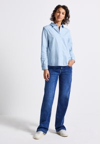 Vrouw die met één hand in haar zak staat, gekleed in een lichtblauw gestreept overhemd, donkerblauwe jeans en witte sneakers, op een effen achtergrond.