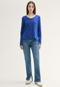 Blauwe gebreide trui met een V-hals, lange mouwen en een losse fit, gecombineerd met lichtblauwe jeans met zijsplitten en witte sneakers.