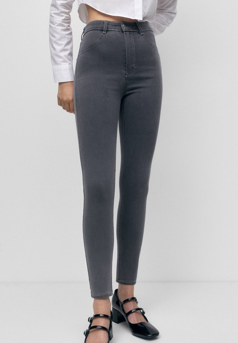 Femme portant un jean skinny gris taille haute, une chemise blanche et des chaussures à talons carrés noires avec une bride à boucle, debout devant un fond clair uni.