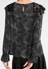 Blusa nera con un motivo testurizzato a pois bianchi, dettagli in pizzo sulle spalle, ruches e un'apertura a chiave sul retro.