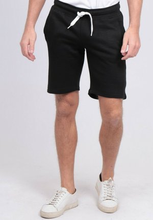 Homme portant un short noir mi-long avec un cordon blanc et des baskets blanches, debout devant un fond uni.
