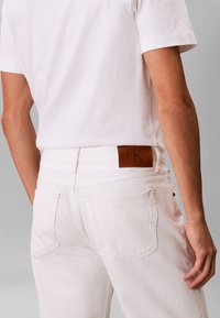 Άνδρας με λευκό τζιν παντελόνι που έχει καφέ δερμάτινο έμβλημα Calvin Klein στη μέση, συνδυασμένο με λευκό κοντομάνικο μπλουζάκι, απεικονίζεται από πίσω.