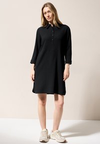 Abito nero con texture, colletto, apertura con bottoni e maniche a tre quarti. Abbinato a scarpe da ginnastica chiare su uno sfondo neutro.
