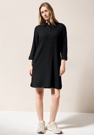 Schwarzes, strukturiertes Kleid mit Kragen, Knopfleiste und Dreiviertelärmeln. Kombiniert mit hellen Sneakers vor einem neutralen Hintergrund.