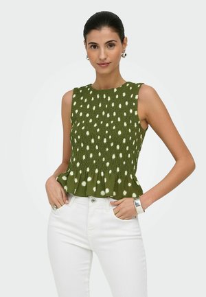 Kobieta nosząca zielony, bezrękawnikowy top z marszczeniami i białymi grochami oraz falbanką na dole, w połączeniu z białymi spodniami, z rękami zgiętymi z dłońmi w kieszeniach.