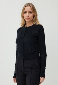 Junge Frau mit blonden Haaren, die eine schwarze knopfne Runde mit floralen Applikationen und schwarze enge Hosen vor einem einfarbigen Hintergrund trägt.