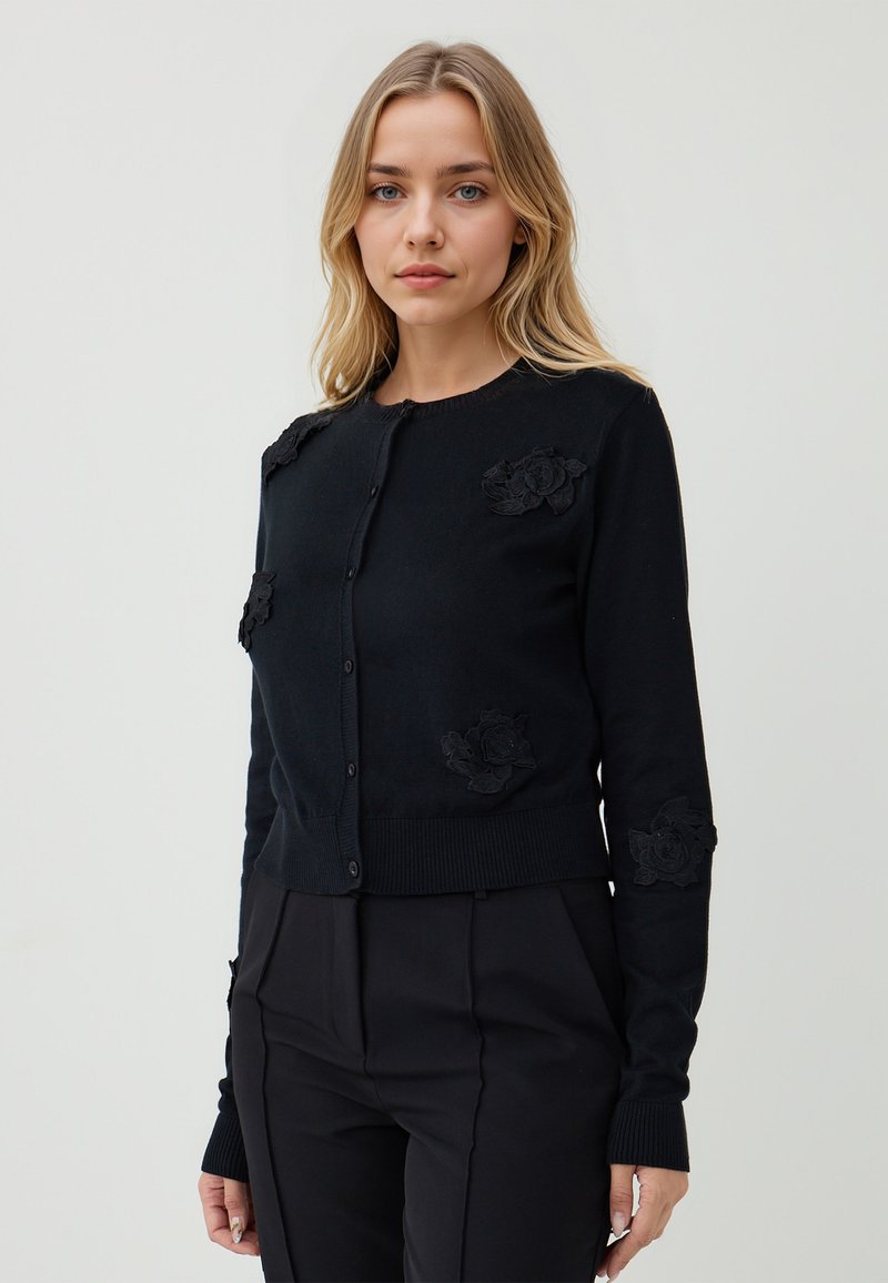 Junge Frau mit blonden Haaren, die eine schwarze knopfne Runde mit floralen Applikationen und schwarze enge Hosen vor einem einfarbigen Hintergrund trägt.