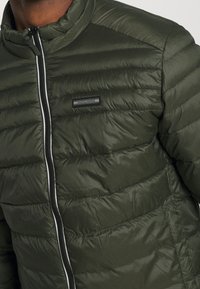 Vadderad olivgrön jacka med ståkrage, dragkedjestängning och kontrasterande silverfärgad dragkedja. Har stickade horisontella linjer och en logotyp.