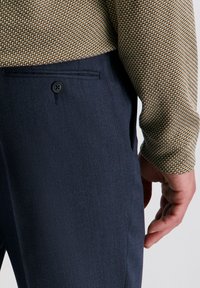 De hand van een man rustend nabij de zak van een marineblauwe broek, terwijl hij een gestreept bruin en beige overhemd met lange mouwen draagt.