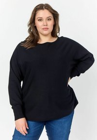 Maglione nero oversize con maniche lunghe, polsini a coste, scollatura rotonda e texture morbida, indossato sopra jeans in denim blu. Design semplice.