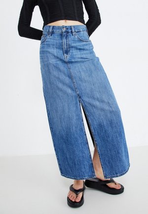 Femme portant une jupe maxi en denim bleu taille moyenne avec une fente à l'avant, un top noir à manches longues et des tongs plateforme noires.