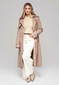 Beżowy trencz z dużymi guzikami, noszony na jasnobeżowym swetrze, w połączeniu z białymi spodniami o szerokich nogawkach i beżowymi butami na spiczastym nosku.