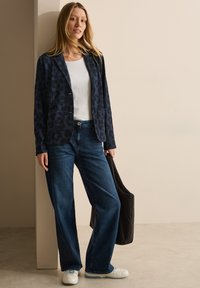 Marineblazer met een luipaardprint, gecombineerd met een witte top en blauwe wijde jeans. Sneakers en een zwarte crossbody tas maken de look compleet.