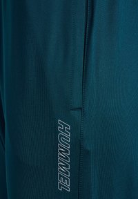 Close-up af mørkegrønblåt stof med lodret søm og hvidt "HUMMEL" logo trykt nær sømmen.