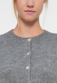 Suéter gris de punto con cuello redondo, textura detallada y tres botones de color claro en la parte frontal.