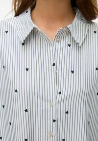 Wit button-up shirt met grijze verticale strepen, versierd met kleine groene hartjes. Het heeft een puntige kraag en beige knopen.