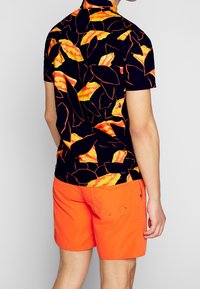 Ein Mann trägt ein schwarzes Kurzarmhemd mit orangefarbenen Blattmustern und leuchtend orangefarbene Shorts, während er vor einem einfarbigen Hintergrund steht.