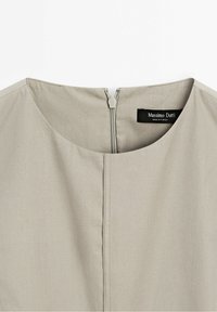Vêtement beige avec un col rond, une couture centrale et une fermeture éclair apparente au dos sous une étiquette noire « Massimo Dutti ».