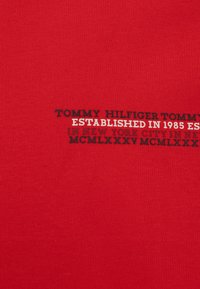 T-shirt in cotone rosso con testo in bianco e nero: "TOMMY HILFIGER FONDATA NEL 1985 A NEW YORK CITY MCMLXXXV MCMLXXX."