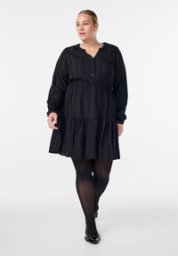 Musta kerroksellinen mekko, joka on valmistettu teksturoidusta kankaasta, jossa on napitettava pääntie ja pitkät hihat, yhdistettynä mustiin sukkahousuihin ja kärjistäviin kenkiin.