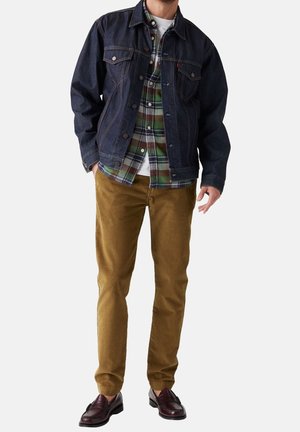 Mann trägt dunkelblaue Jeansjacke über grün-braun kariertem Hemd, braune Hose und dunkelbraune Lederloafers auf weißem Hintergrund.