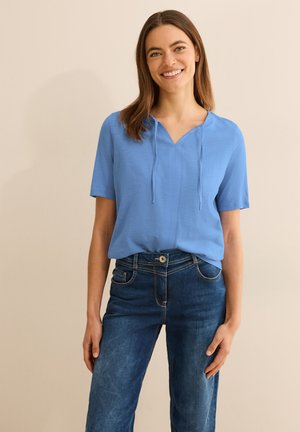 Haut bleu à manches courtes avec décolleté en V et détail à cordon de serrage, associé à un jean bleu foncé avec poches avant et couture contrastante.
