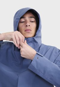 Chaqueta impermeable de color azul claro con capucha, cuello abrochado y puños con botones. Textura suave y detalles de diseño mínimos.