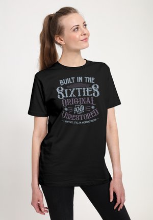 Ung kvinde med hestehale iført en sort T-shirt med teksten "Built in the Sixties Original and Unrestored", stående foran en ensfarvet baggrund.