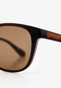 Bruntonede solbriller med en blank sort ramme. Stangen har orange "SUPERDRY" branding, der fremhæver et moderne design.