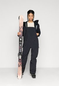 Σαπούνι σκι που περιλαμβάνει μαύρα μόνωτα σαλοπέτα, ανοιχτό μπεζ μπλούζα και μαύρη ζακέτα. Το μοντέλο κρατάει ροζ και μαύρα σκι με δεσίματα. Μαύρα γυαλιά σκι.