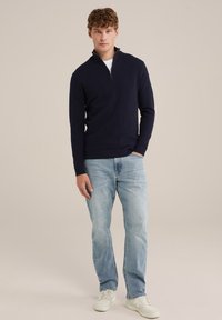 Marinefarbener Reißverschluss-Pullover aus leichtem Strick, kombiniert mit hellblauen Jeans und weißen Sneakers. Das Model steht vor einem neutralen Hintergrund.