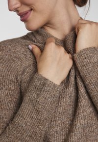 Brauner gestrickter Pullover mit hohem Kragen, der eine strukturierte Oberfläche und gerippte Bündchen aufweist. Hände halten den Stoff nahe am Kragen.