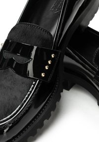 Schwarzer Lacklederloafer mit einem Obermaterial mit Fellstruktur, goldenen Nieten am Riemen und robustem Gummisohle auf weißem Hintergrund.