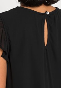 Donna che indossa una blusa nera con maniche corte a volant e una piccola apertura a forma di chiave sul retro, chiusa da un bottone bianco.