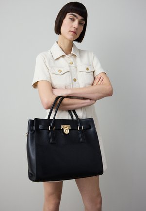 Sort lærhåndveske med gullfarget metall, rektangulær form, to håndtak på toppen og en låsdetalj, plassert mot en lys bakgrunn.