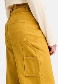Gula cargobyxor gjorda av slitstark tyg, med flera fickor, sydda detaljer och en midjebandsetikett med Timberland-logotypen.