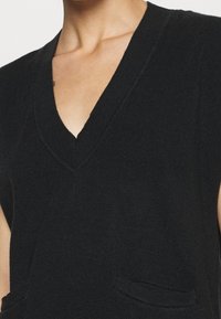 Svart ärmlös stickad topp med V-ringning och två framfickor, med en ribbad textur och en avslappnad passform.