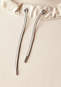 Beżowa bluzka z marszczonym dekoltem i sznurkiem. Sznurek posiada pleciony wzór z metalowymi końcówkami. Gładka faktura materiału.