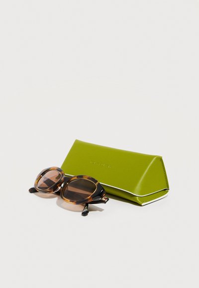 Lunettes de soleil rondes en écaille de tortue avec des verres bruns à côté d'un étui à lunettes en cuir vert vif fermé sur une surface blanche.
