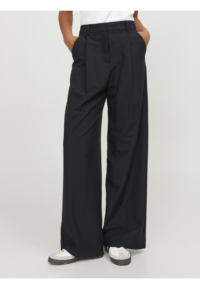 Pantalon noir droit à jambes larges porté avec des baskets blanches et un top blanc rentré, les mains dans les poches.