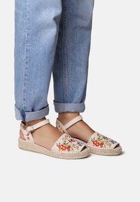 Beige espadrilles with floral embroidery in red, yellow, and blue. Features a woven jute sole and an ankle strap. Worn with cuffed denim jeans.