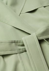 Světlá zelená látka s propracovaným prošitím a uvázaným páskem, pravděpodobně součást oděvu, jako je kabát nebo šaty.